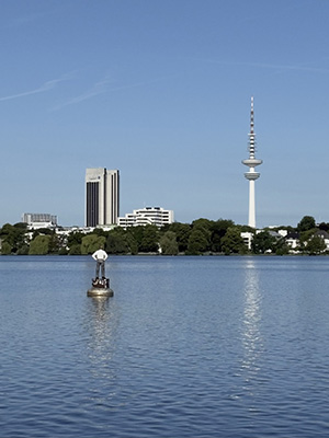 City skyline with a tall tower and buildings across a lake, featuring a small statue in the water.